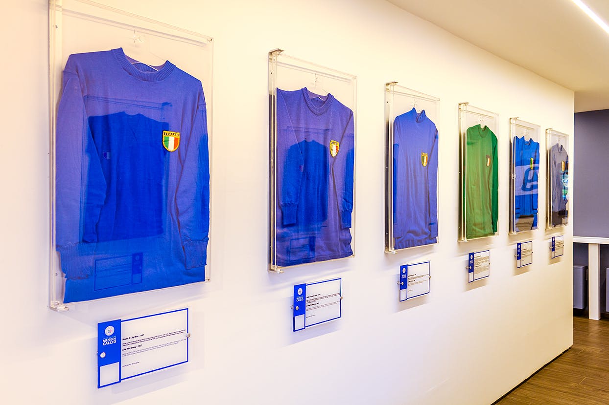 Fyra inramade vintage fotbollströjor, inklusive tre blå och en grön, visas på en vägg med beskrivande plakat under dem.