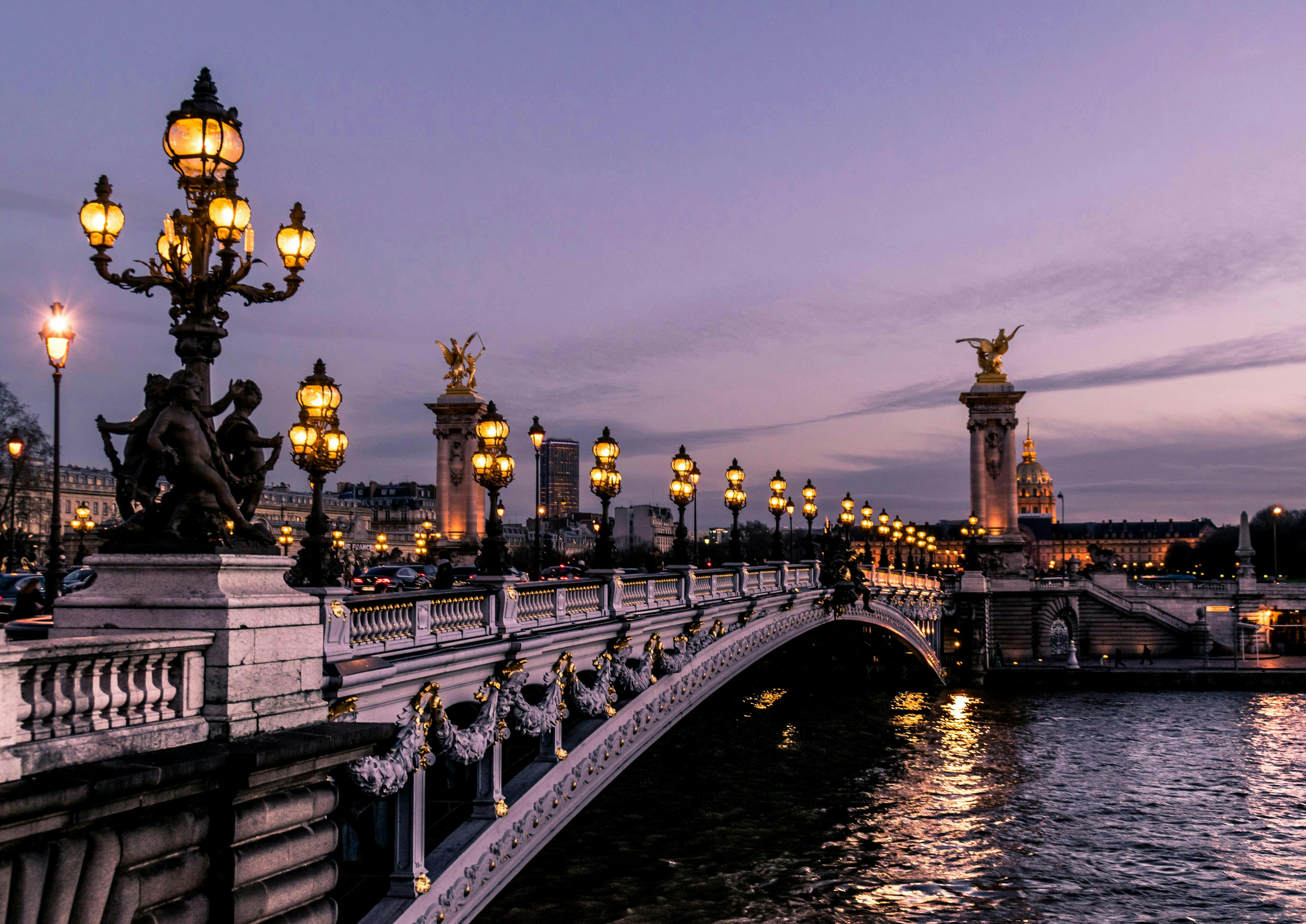 A bridge adorned with ornate streetlights and statues at twilight, with a cityscape and a golden-domed building in the background.