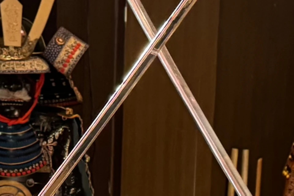 Two individuals in traditional attire practicing a sword duel in a room with samurai armor displayed in the background.