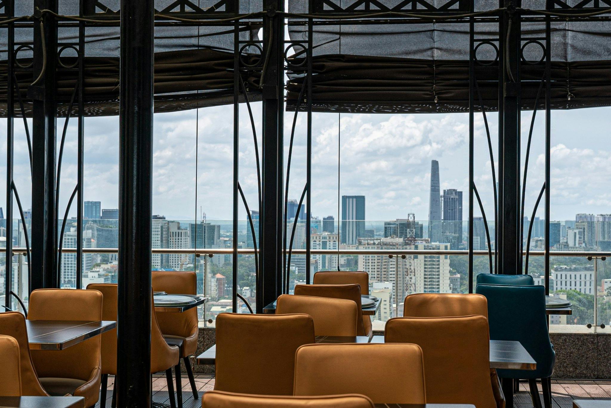 A restaurant with brown leather chairs and glass tables, overlooking a cityscape with tall buildings under a cloudy sky.