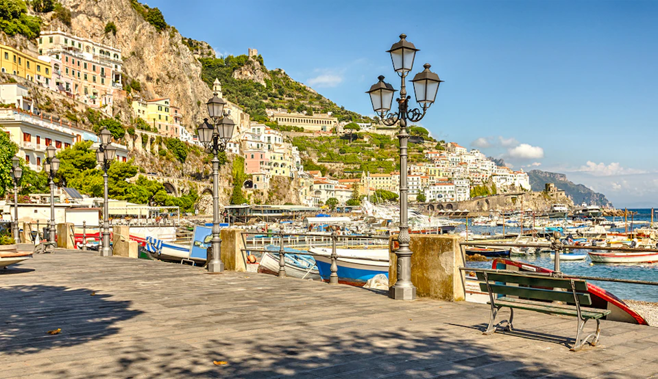 Seaside promenade with streetlamps, benches, and boats docked in a harbor. Hillside buildings and green cliffs in the background.
