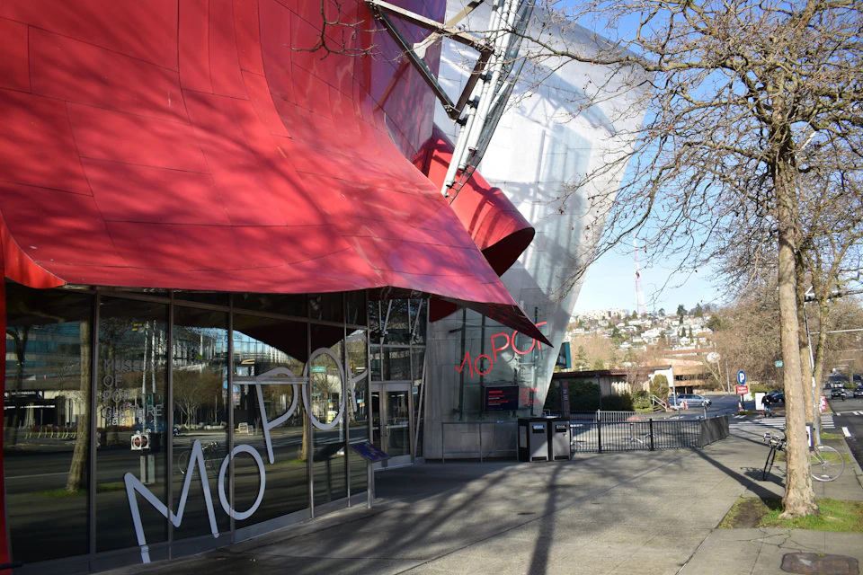 Red and silver modern building with large windows, bold "MoPOP" letters, and bare trees. Urban backdrop with hilly terrain.