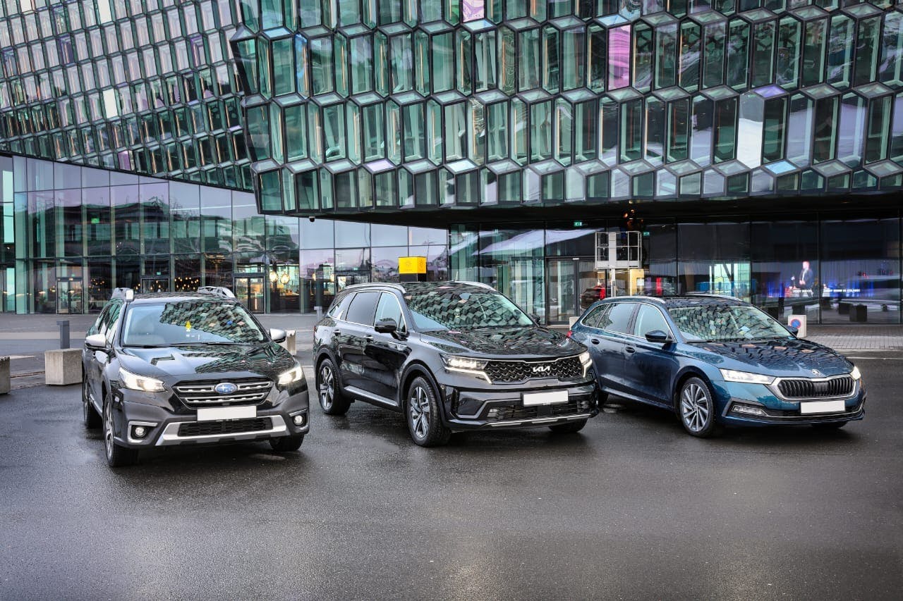 Three cars—a black Subaru, a black Kia, and a blue Skoda—parked on a wet pavement in front of a modern glass building.