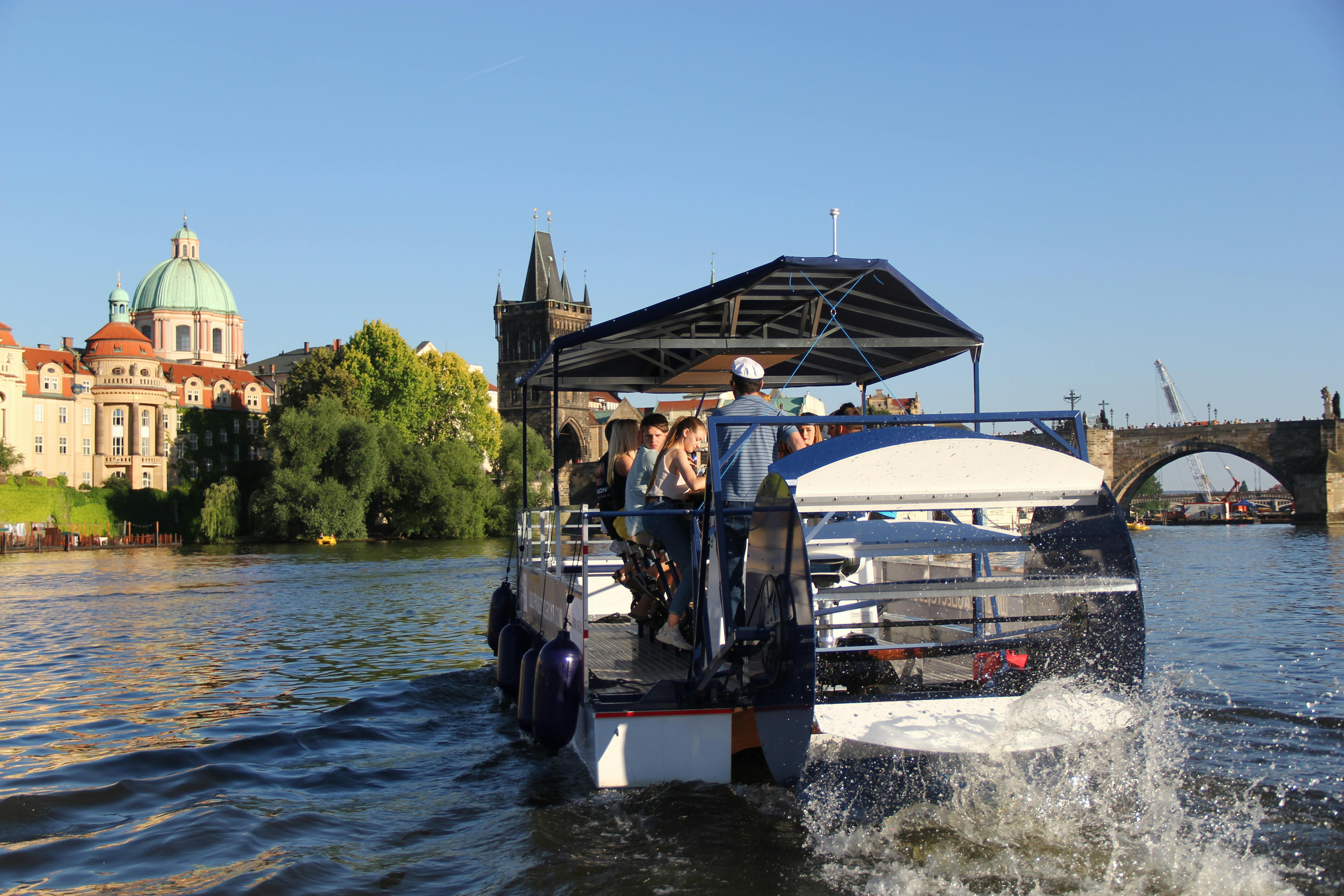 La gent viatja en un vaixell cobert de dosel en un riu, amb edificis històrics i una torre alta visible al fons sota un cel blau clar.