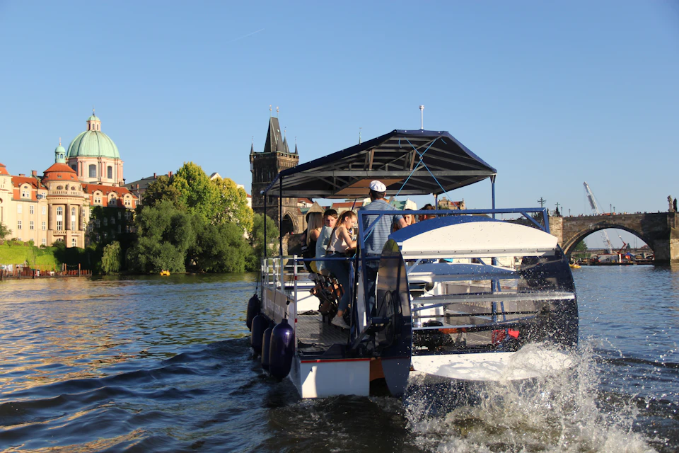 People ride on a canopy-covered boat on a river, with historic buildings and a tall tower visible in the background under a clear blue sky.