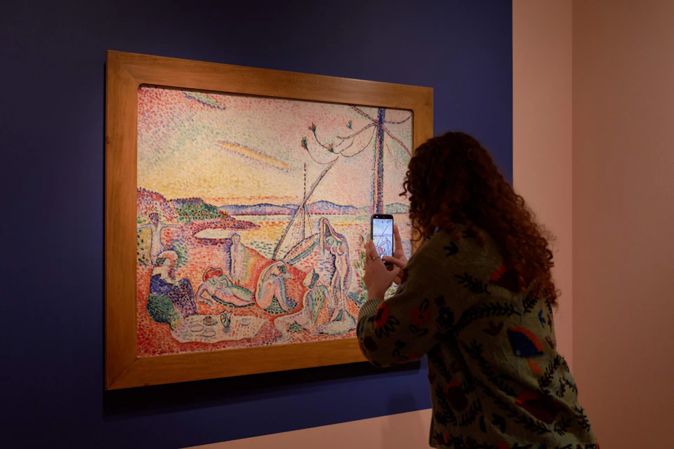 A person photographing a framed pointillist painting displayed on a blue wall in a gallery.