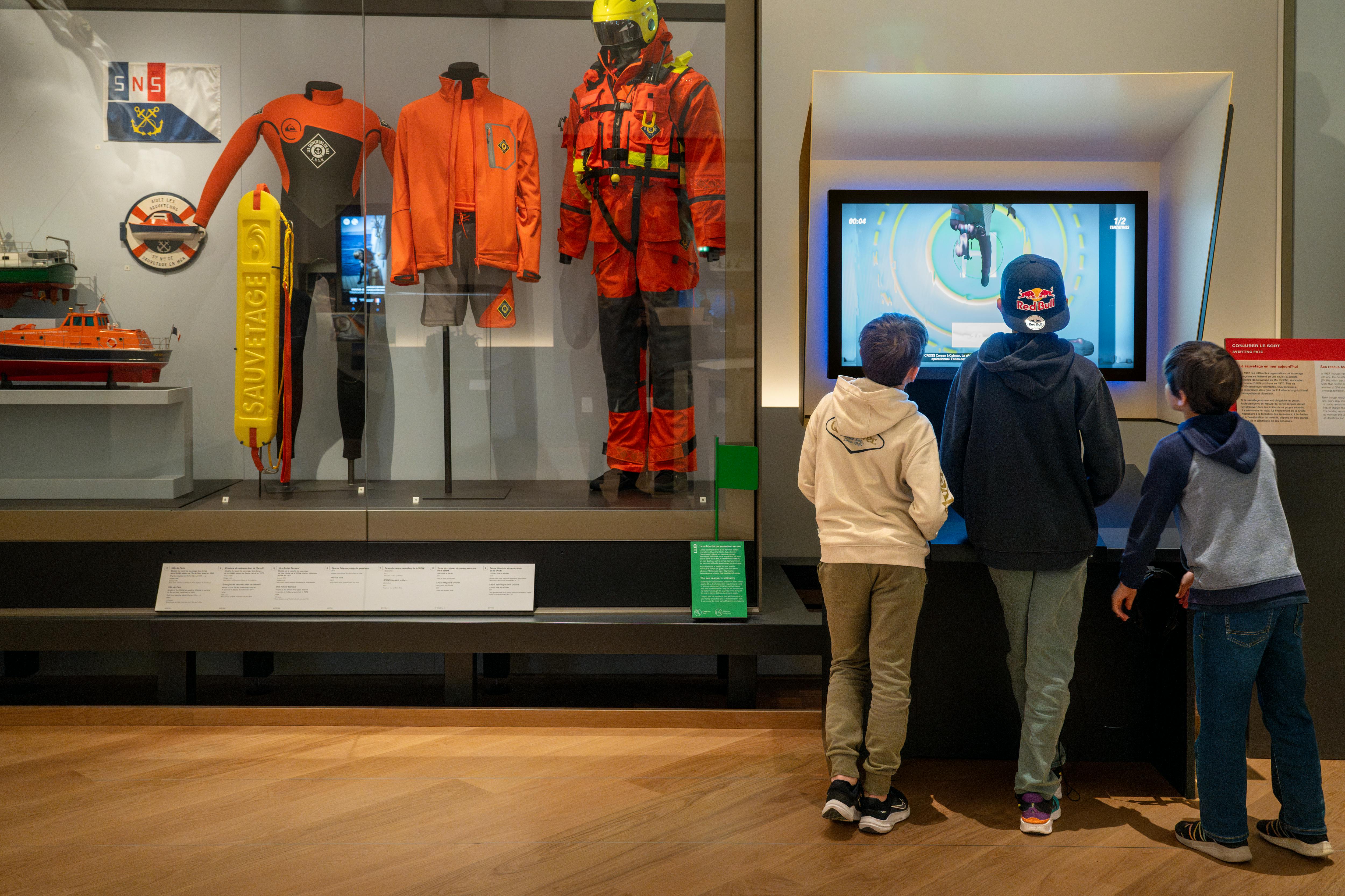 Tre barn tittar på en interaktiv skärm i ett museum, med räddningsuniformer och livbåtsmodeller som visas i lådor i närheten.