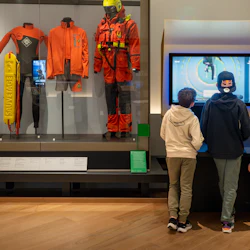 Tre barn tittar på en interaktiv skärm i ett museum, med räddningsuniformer och livbåtsmodeller som visas i lådor i närheten.