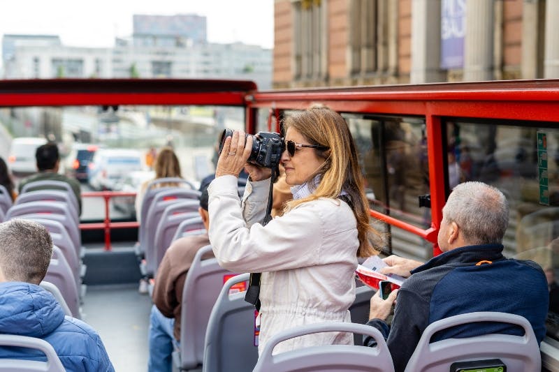 Une femme prend une photo avec un appareil photo dans un bus rouge à toit ouvert tandis qu'une autre personne lit une brochure.