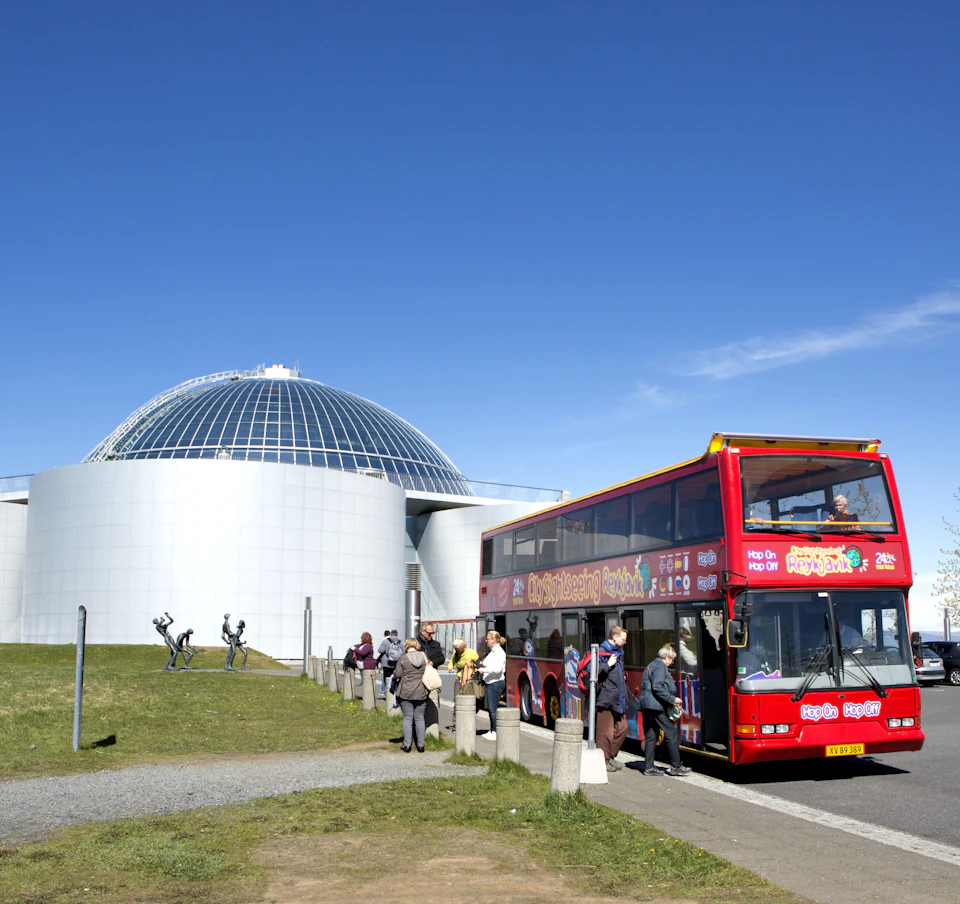 A red double-decker tour bus with passengers is parked beside a modern, dome-shaped building under a clear blue sky.