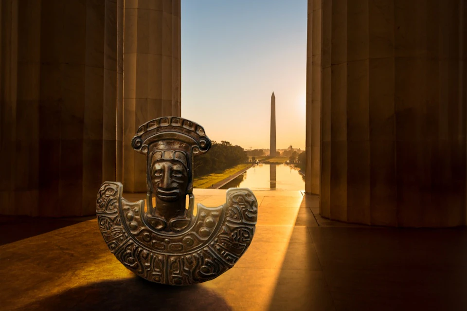 Ancient sculpture in foreground with view of Washington Monument and Reflecting Pool at sunset between pillars.