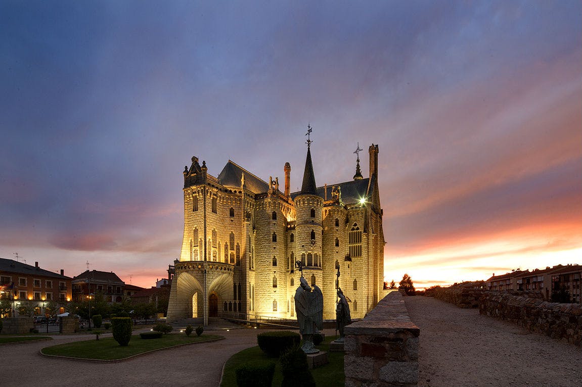 A lit-up castle with Gothic architecture at sunset, featuring arched windows, spires, statues, and a garden pathway.