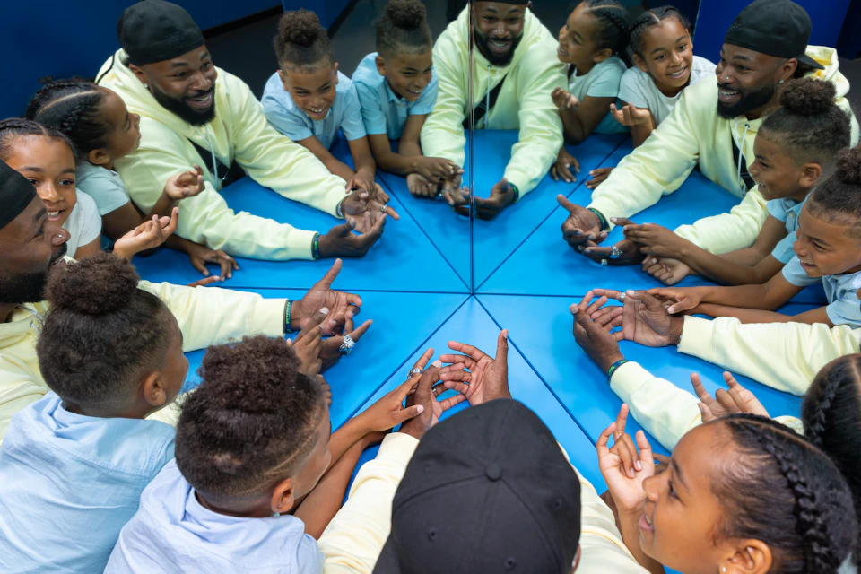 A group of children and an adult interact at a mirrored table, their reflections creating a symmetrical effect.