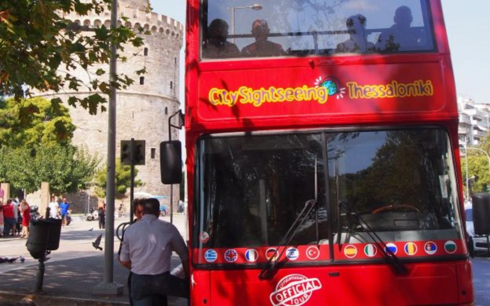 Red double-decker tour bus near a historic stone tower. People visible in the bus's upper deck, shadows of trees overlay the bus.