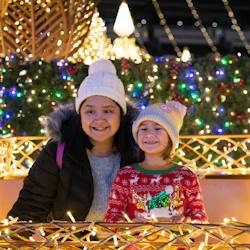 Two people wearing winter attire smiling in a festive, illuminated setting with decorative lights and greenery.