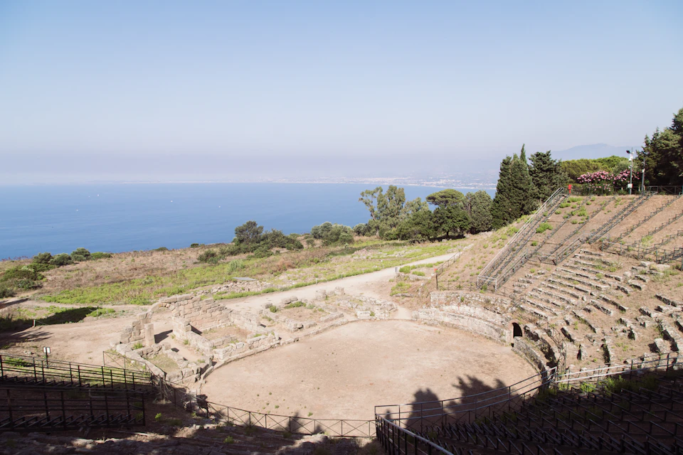 Ancient amphitheater with stone seating and open circular stage, overlooking a coastal landscape with trees and a clear blue sky.