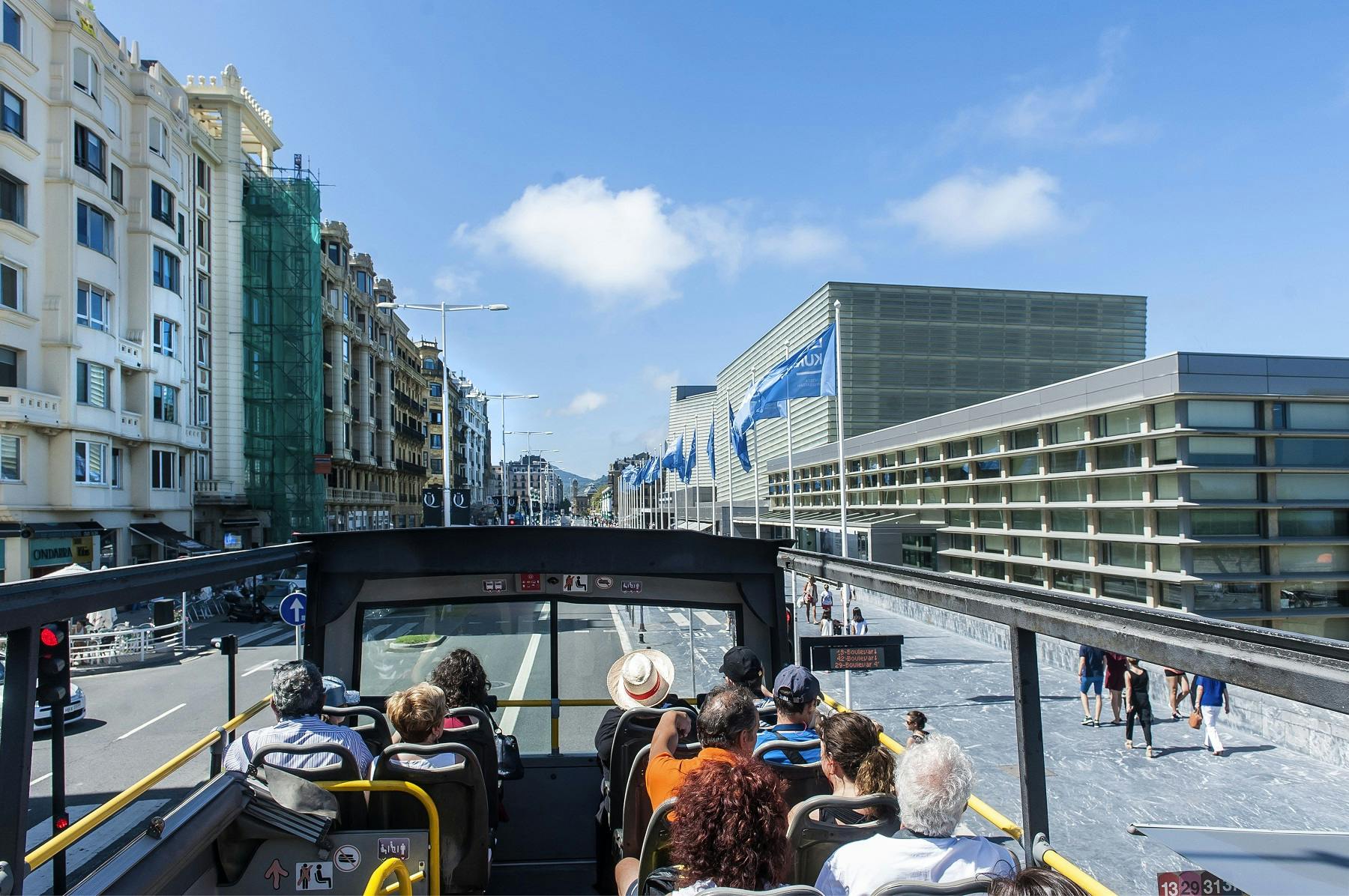 A red double-decker tourist bus in San Sebastián, with various icon labels, is parked near a historic building.