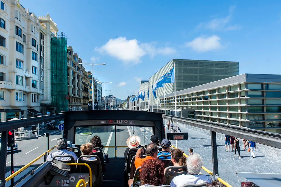 A red double-decker tourist bus in San Sebastián, with various icon labels, is parked near a historic building.