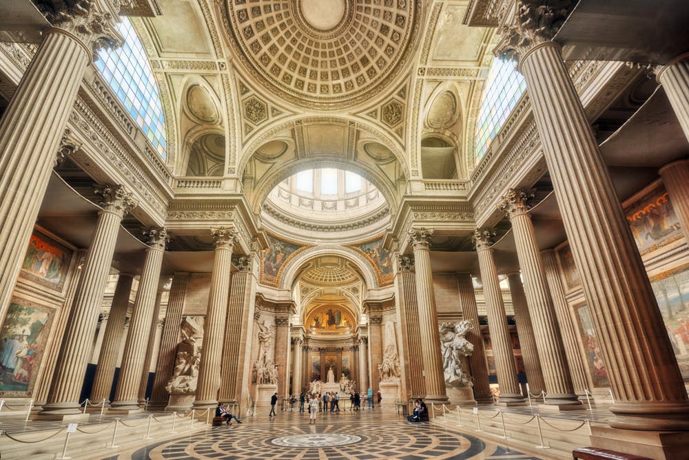 A grand hall with an ornate domed ceiling, large columns, statues, stained glass windows, and people walking on patterned floors.