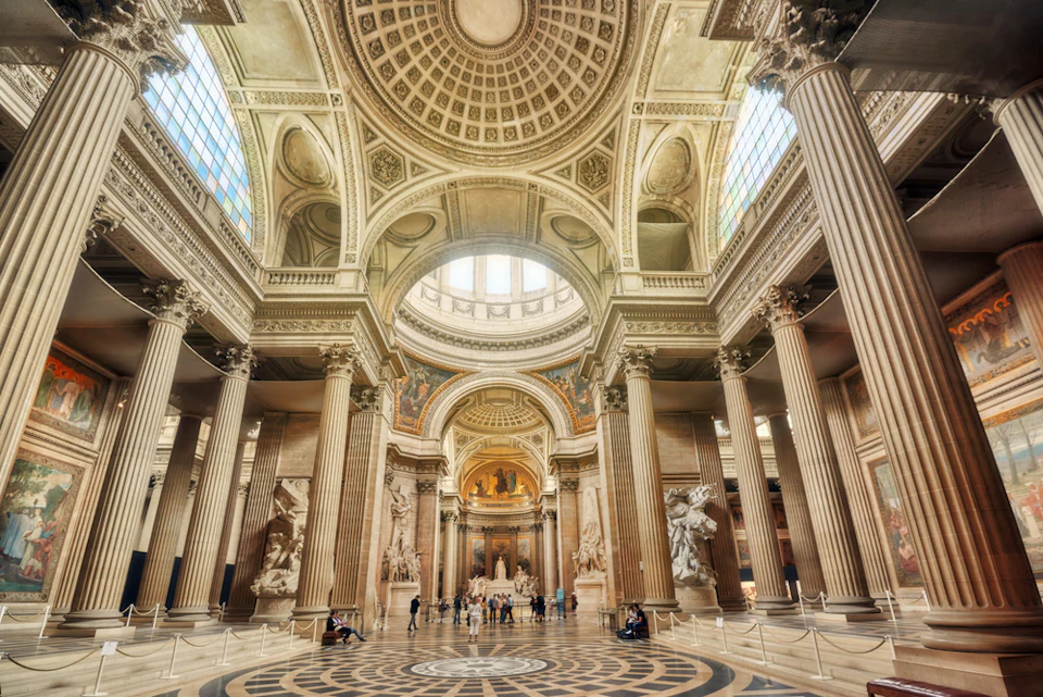A grand hall with an ornate domed ceiling, large columns, statues, stained glass windows, and people walking on patterned floors.