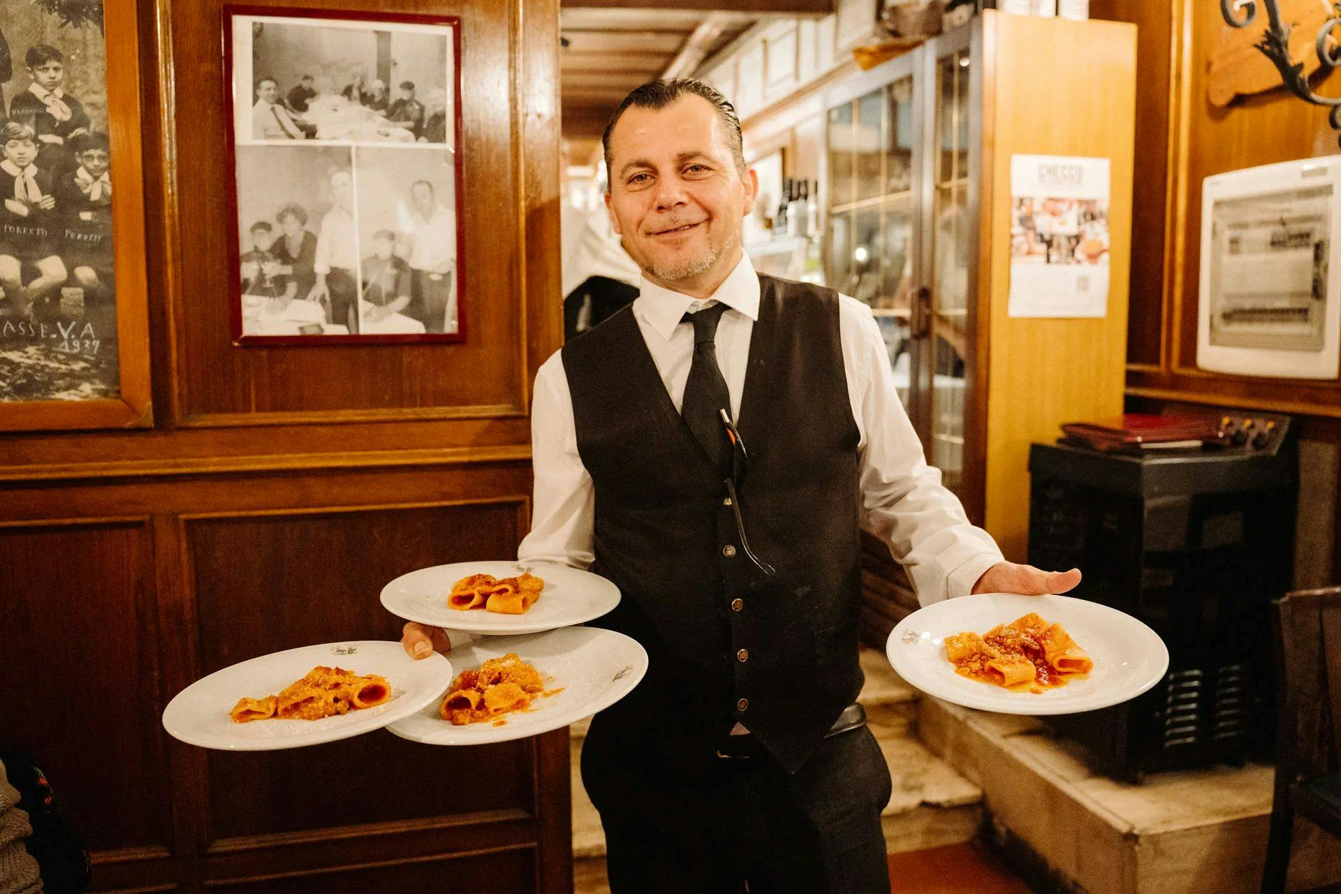 A waiter in a white shirt and black vest smiles while holding four plates of pasta in a warmly lit restaurant.