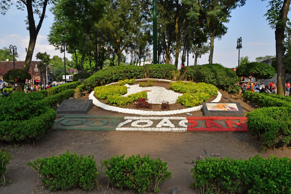 Landscaped garden with plants arranged to form a circular design. "COYOACÁN" spelled out in colored rocks at the front.