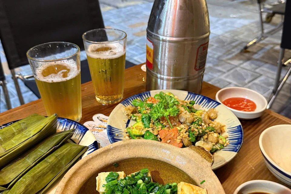 A table with two glasses of beer, a plate of mixed stir-fry, a pot of tofu soup, wrapped leaf parcels, and a metal beverage container.