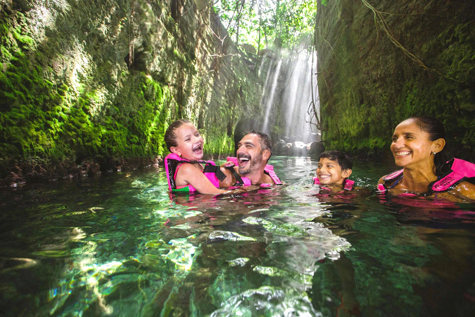 A family of four wearing life jackets smiles and enjoys swimming in a green canyon with sunlight streaming through trees above.