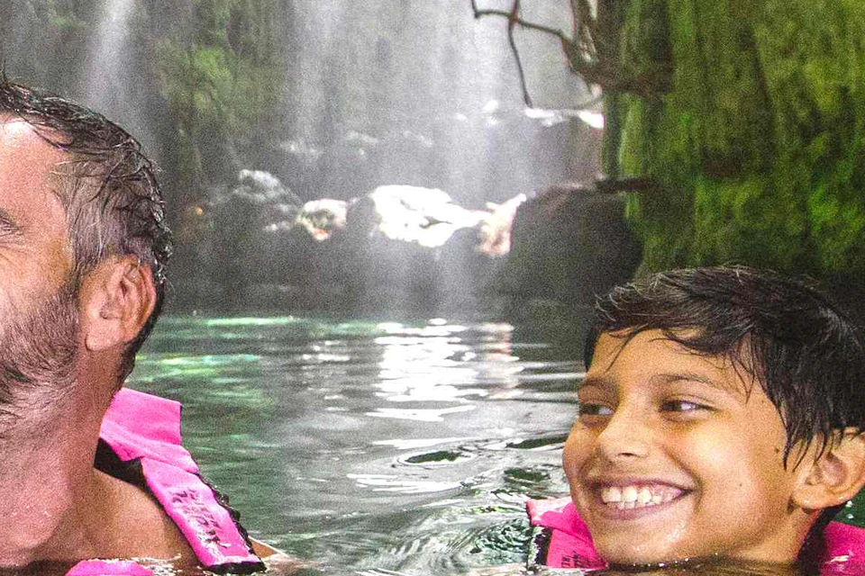 A family of four wearing life jackets smiles and enjoys swimming in a green canyon with sunlight streaming through trees above.