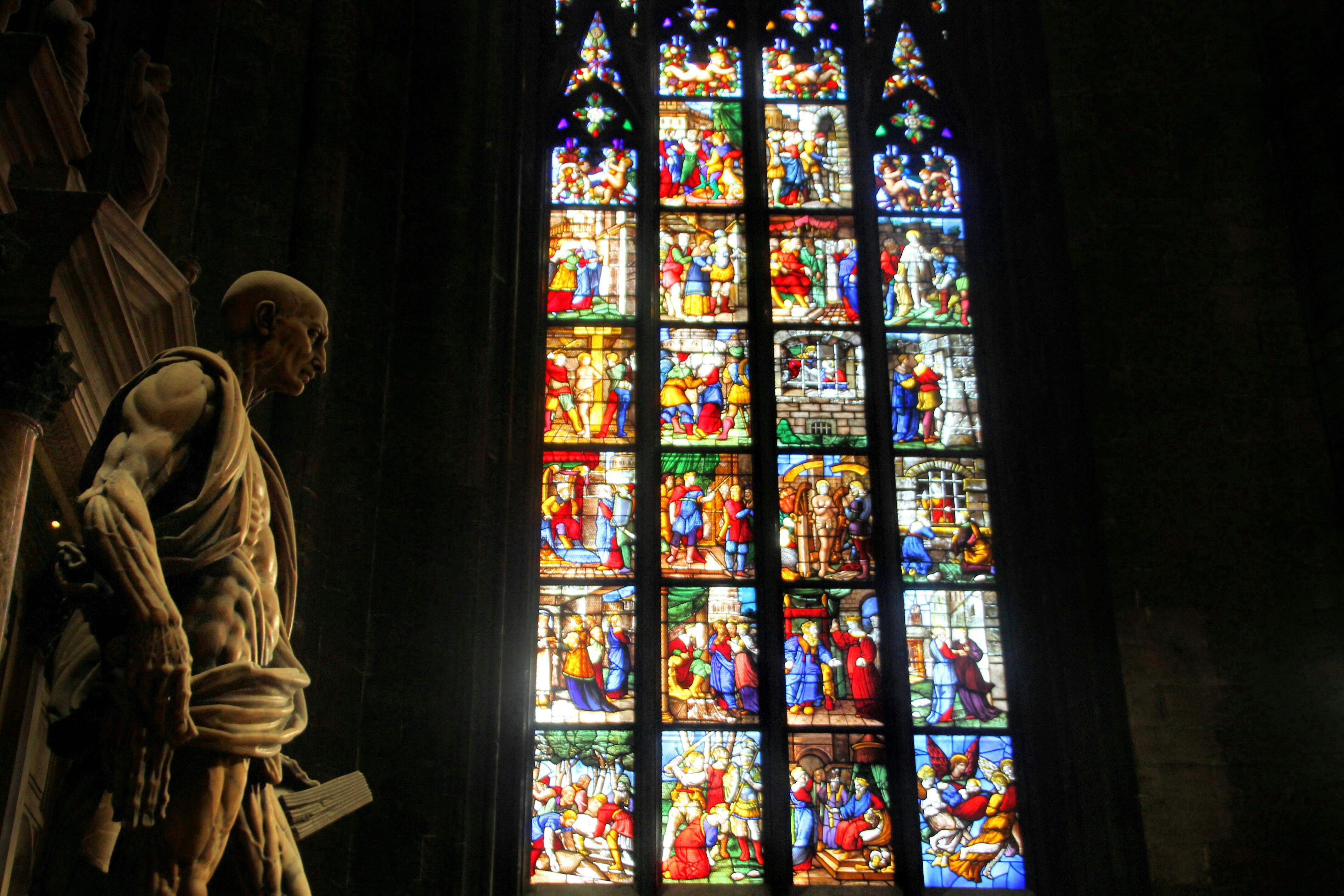 A statue stands beside a large, intricate stained glass window depicting various colorful religious scenes.