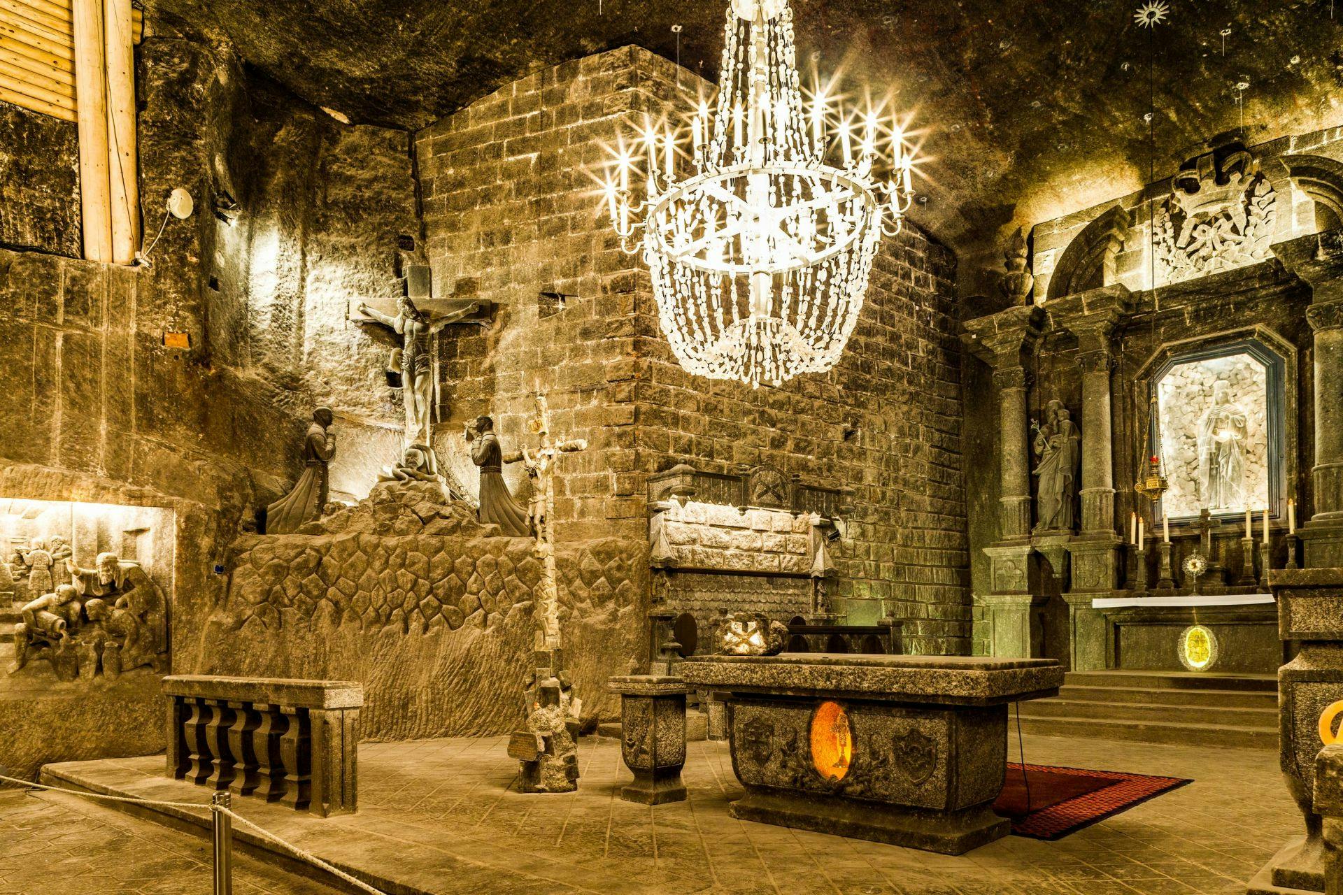 A dimly lit underground chapel with stone walls, a large crystal chandelier, and religious sculptures and altar.