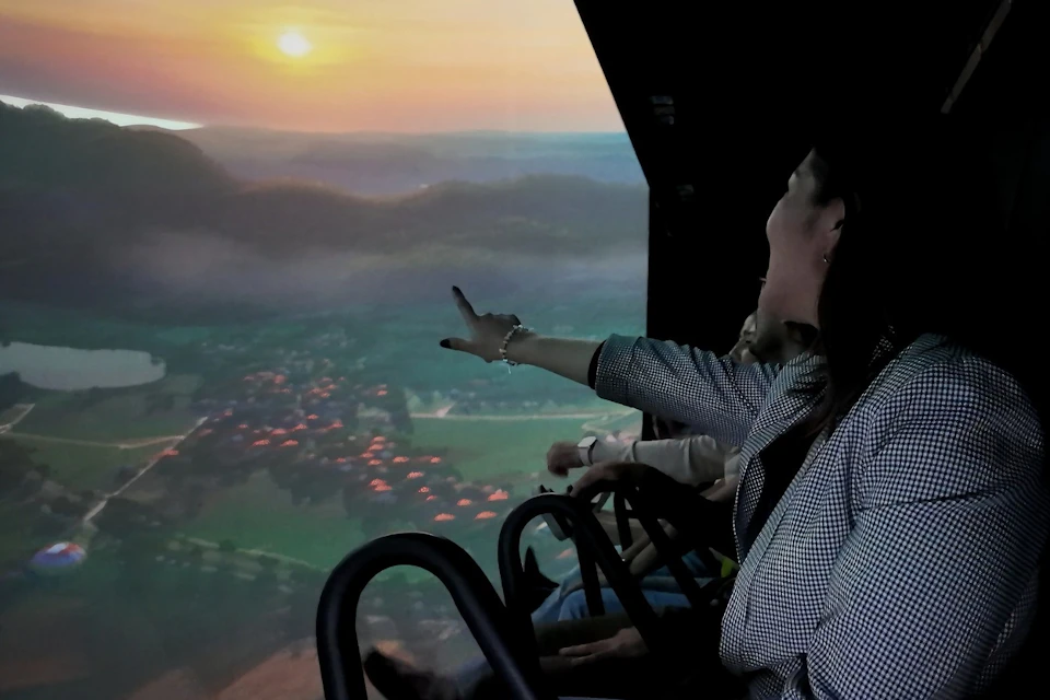 People seated on a ride with a panoramic screen, showing a scenic landscape at sunset. A woman points towards the screen.