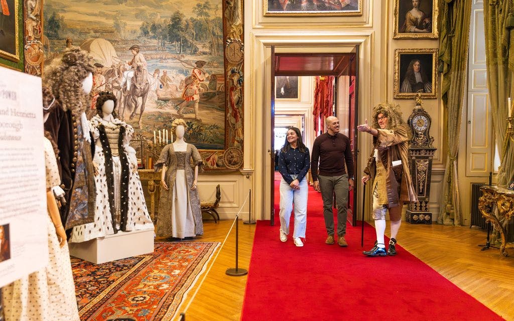 一位身着时代服装的导游带领两位游客穿过一间华丽的房间，里面有挂毯、绘画和历史悠久的装饰。