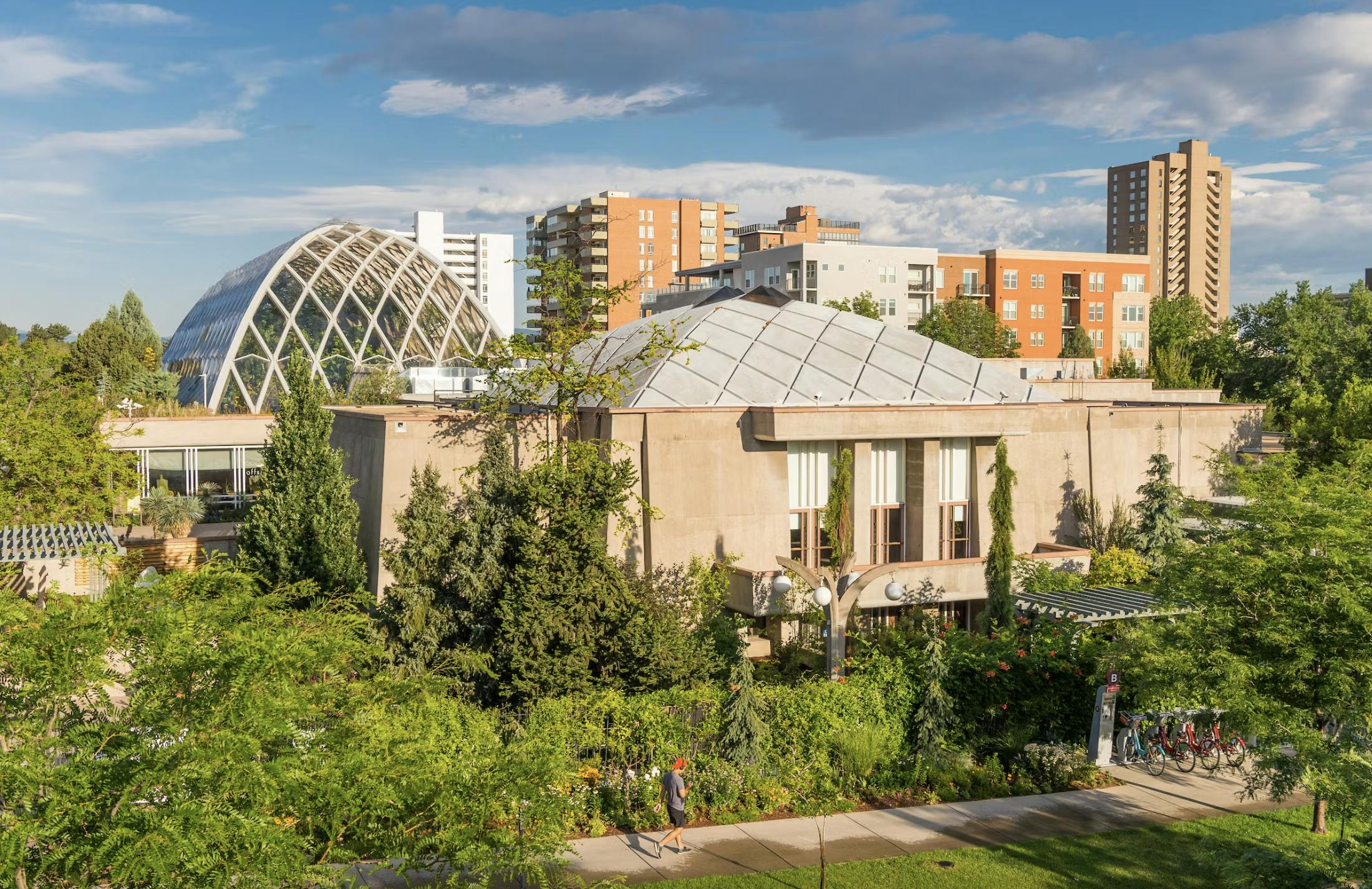 Een groen park met gebouwen, waaronder een koepel en een hoekige structuur, omringd door bomen en voetpaden op een zonnige dag.