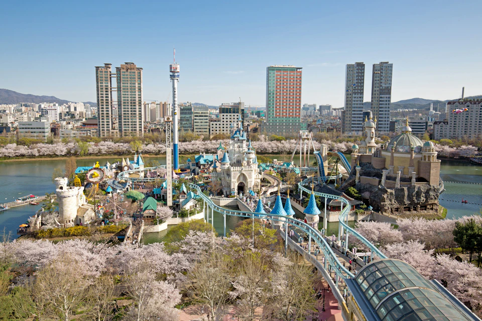 Aerial view of an amusement park featuring a castle, rides, and cherry blossom trees, with a city skyline in the background.