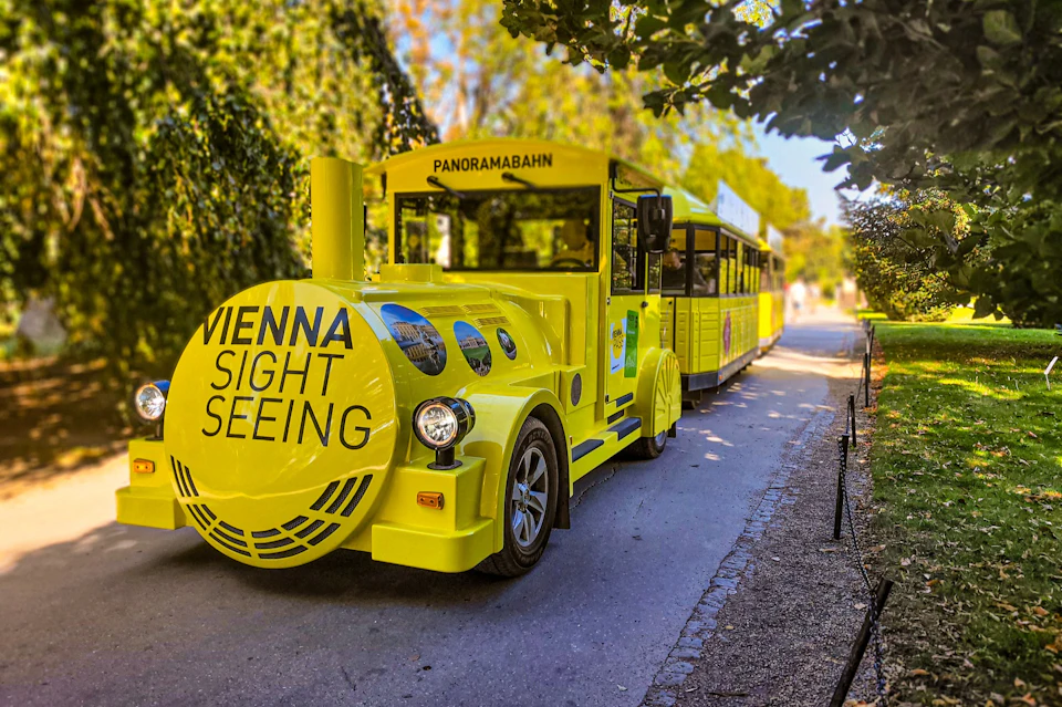 A yellow sightseeing train labeled "Vienna Sightseeing" on a park pathway, surrounded by greenery.