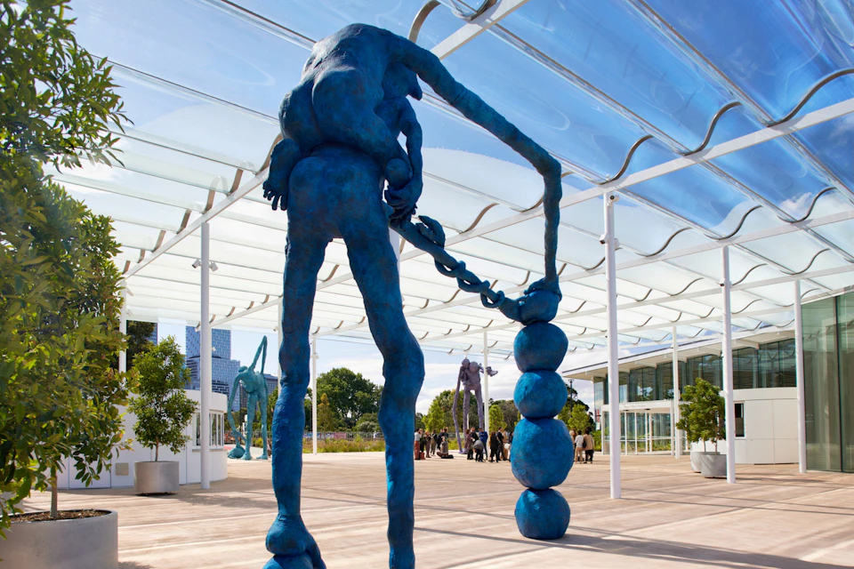 Blue abstract sculpture of a figure with a chain-like object, set in an outdoor space with a glass canopy and people in the background.