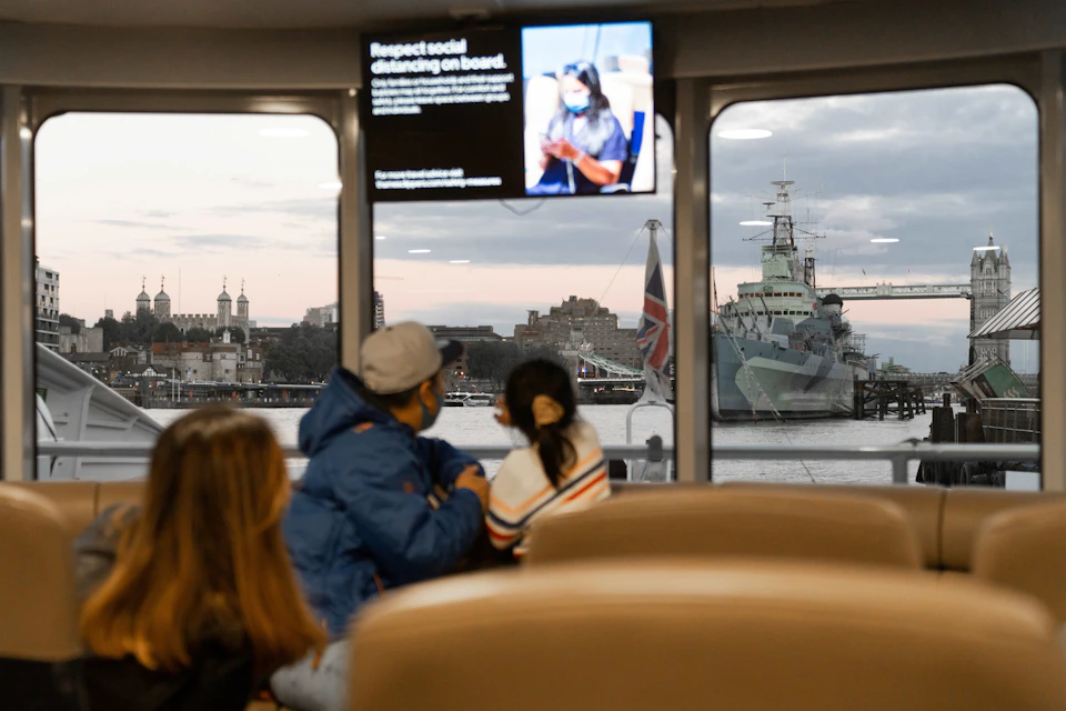 Passengers seated indoors on a ferry, looking out at a harbor with a large ship and buildings visible through windows.