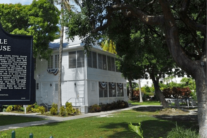 A white, two-story house with patriotic bunting is surrounded by trees and a well-maintained lawn under a blue sky.