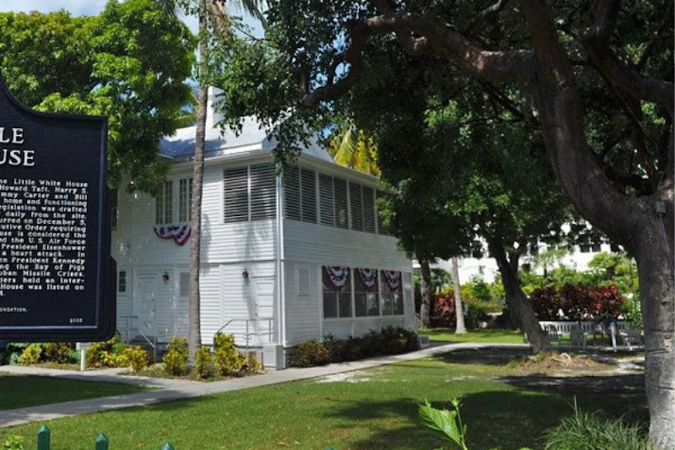 A white, two-story house with patriotic bunting is surrounded by trees and a well-maintained lawn under a blue sky.