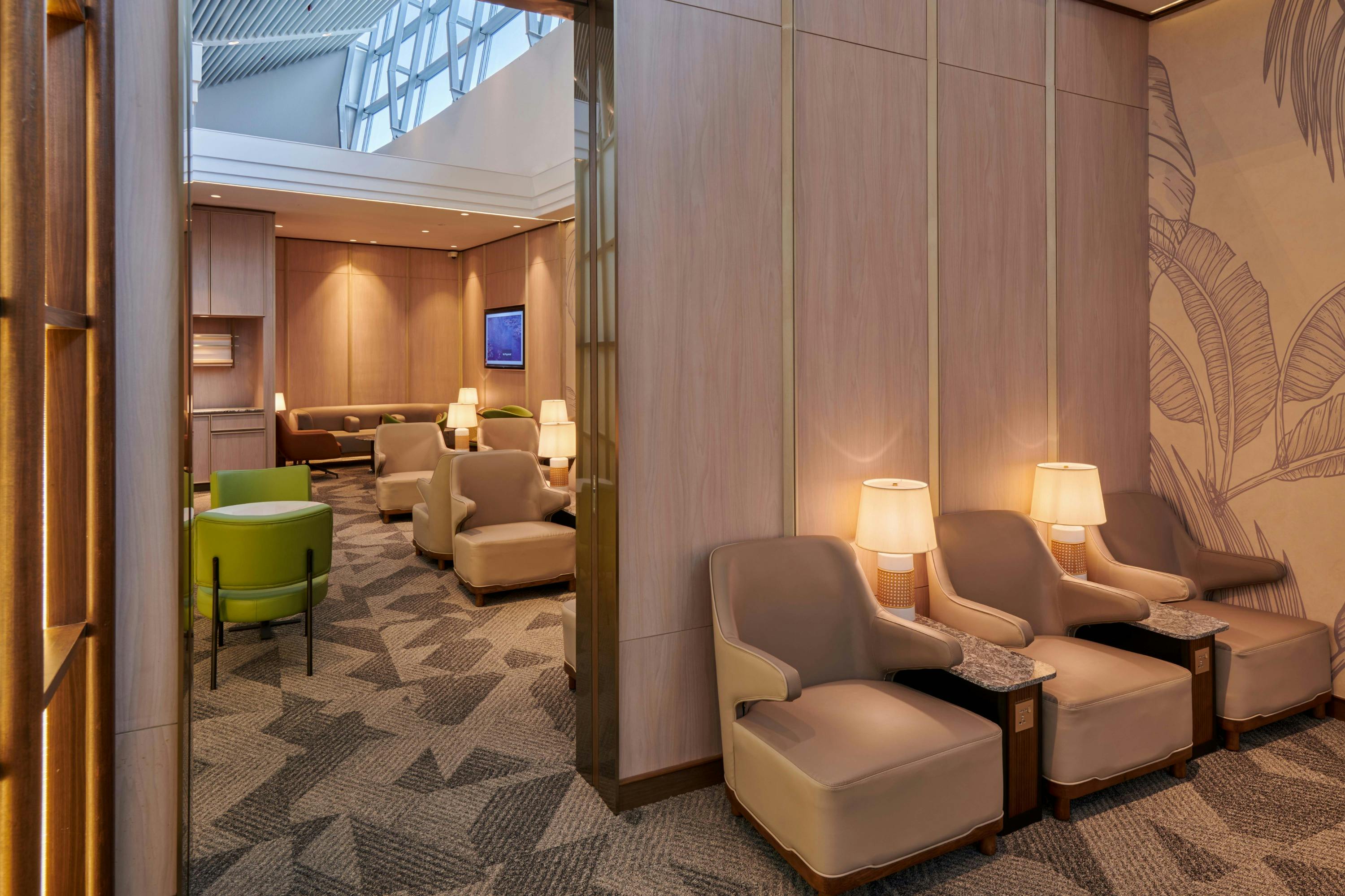 A stylish lounge interior with beige armchairs, modern lighting, green seating, and patterned carpeting under a high ceiling.