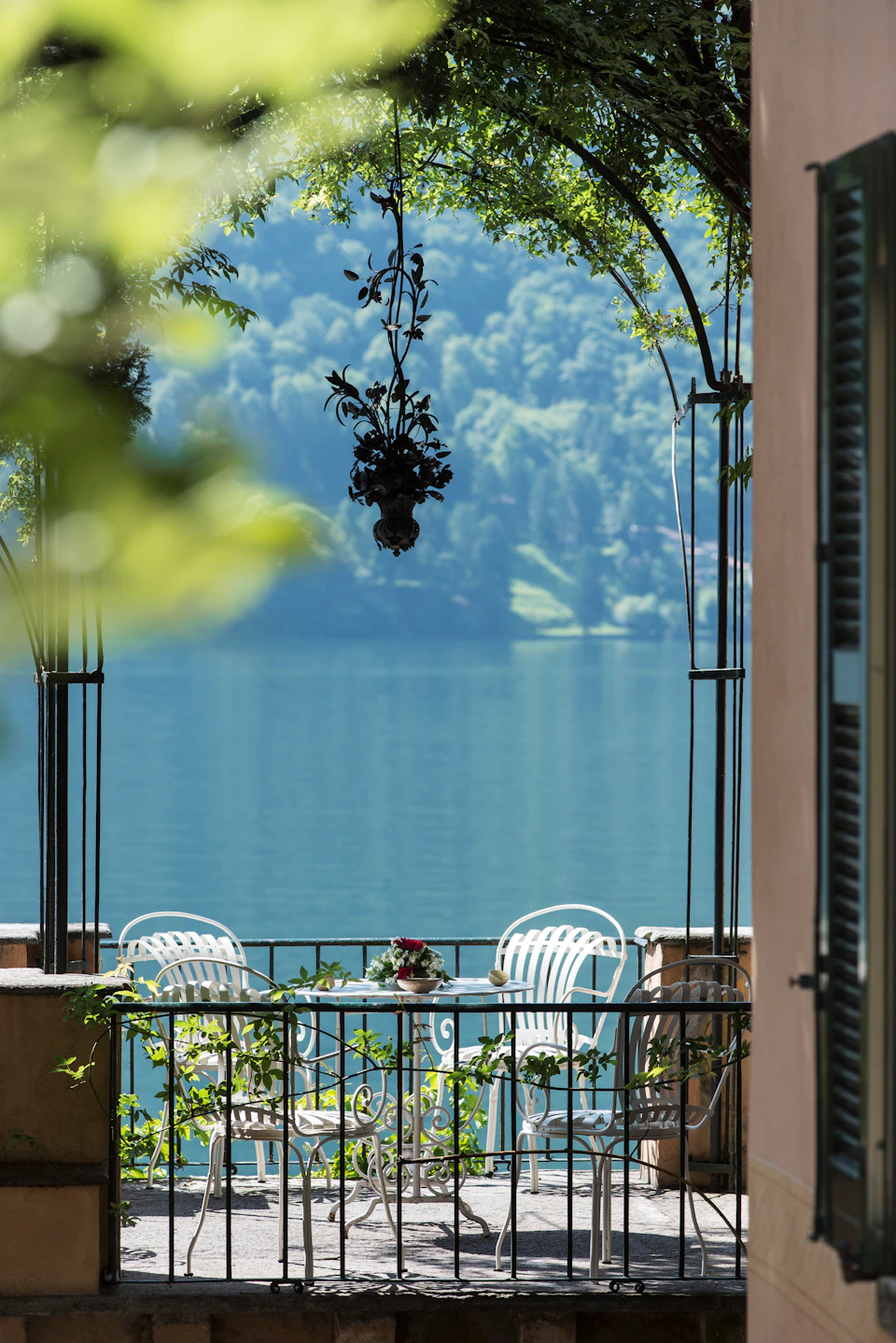 A lakeside patio with a round table surrounded by white chairs, featuring a floral centerpiece and hanging plant, with trees visible in the background.