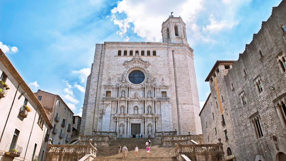 Large stone cathedral with ornate facade, wide staircase, and several people ascending. Sky is bright with scattered clouds.