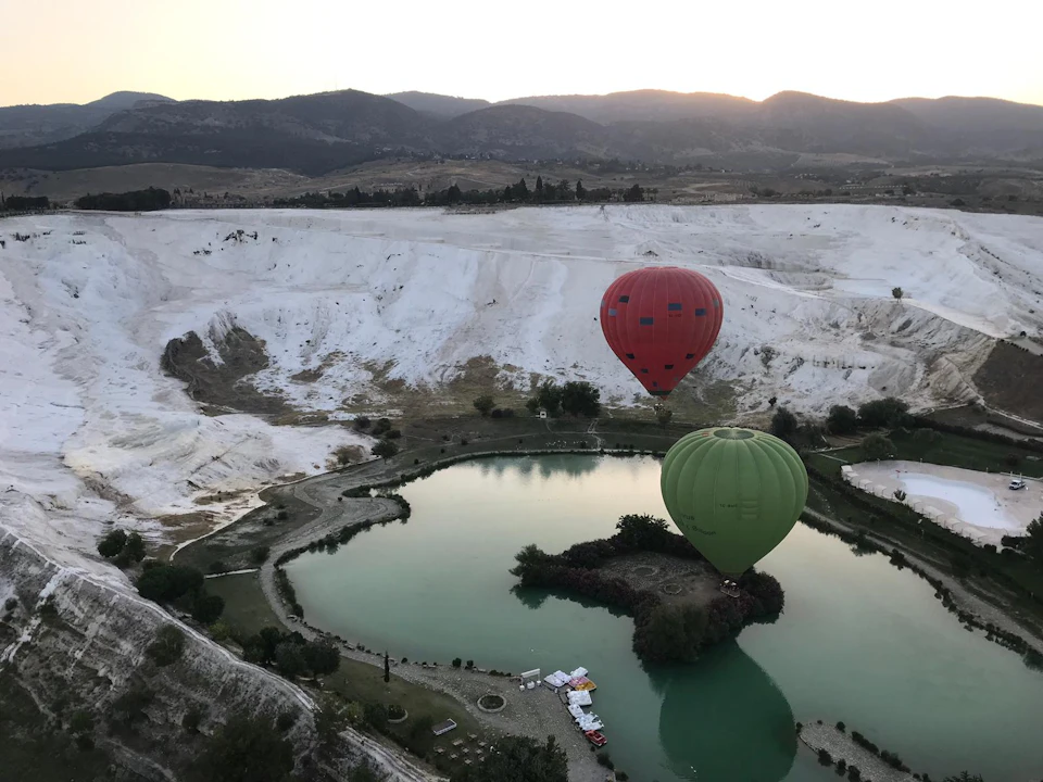 Two hot air balloons, one red and one green, floating over a serene lake surrounded by white rock formations at sunrise.