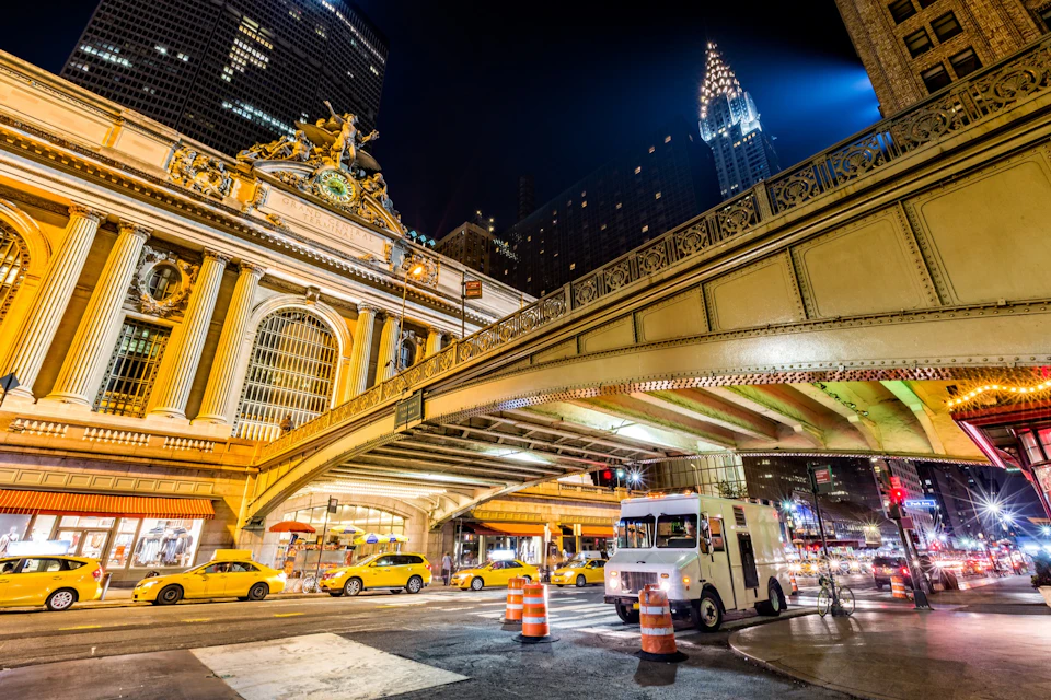 Night scene of Grand Central Terminal with yellow taxis and a white truck, under an overpass with the Chrysler Building in view.