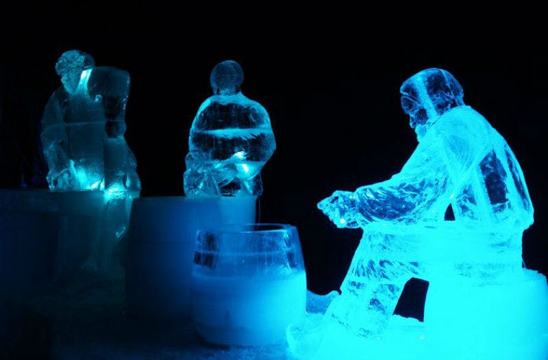 Three ice sculptures of people sitting and standing around ice barrels, illuminated by blue lights against a dark background.