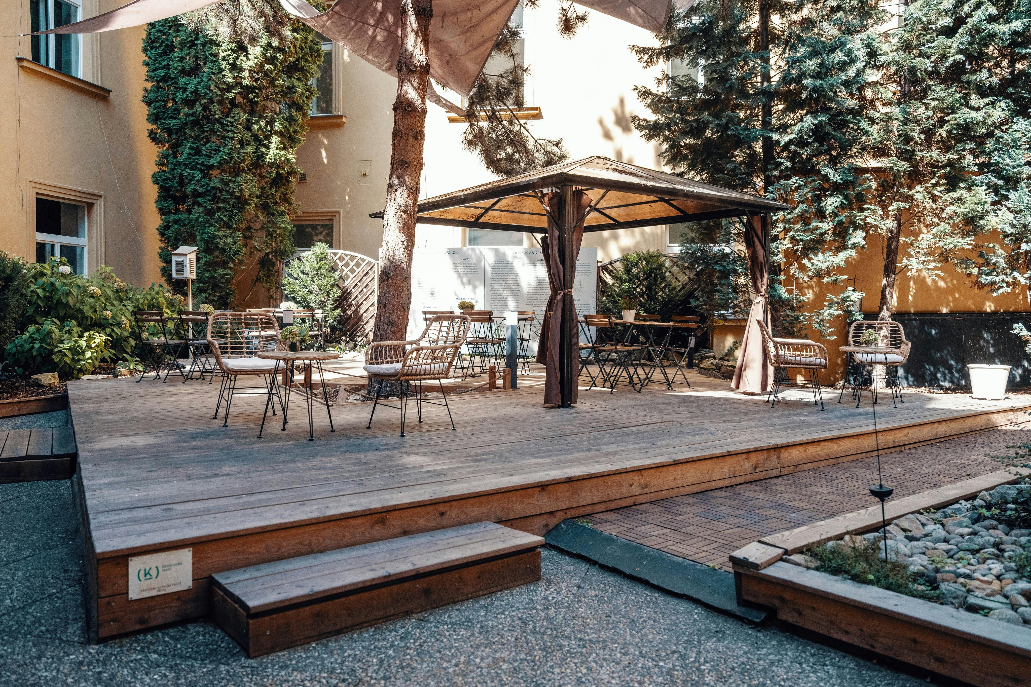 Outdoor patio with wooden deck, empty tables and chairs, shaded by a canopy, surrounded by trees and plants.