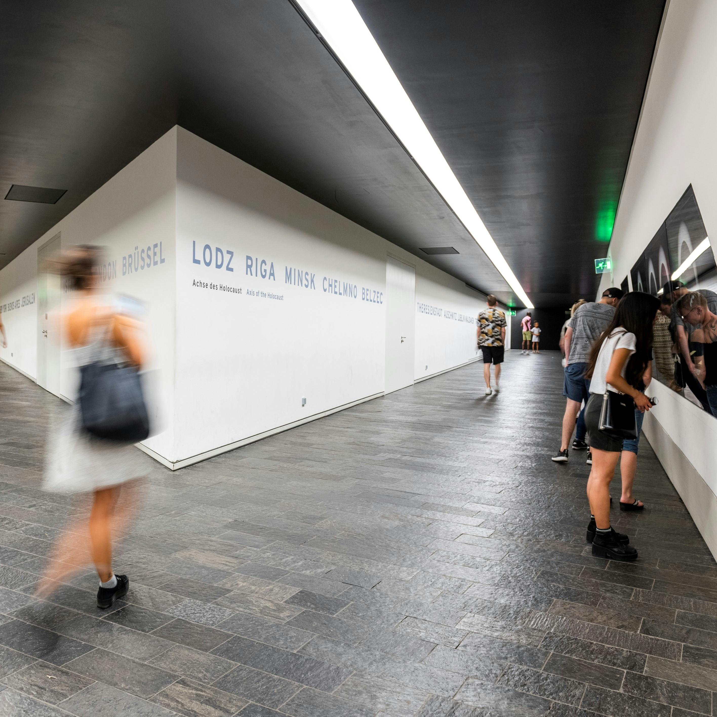 Lidé procházející moderní, dobře osvětlenou chodbou s bílými zdmi zobrazujícími názvy měst v modré barvě. Někteří lidé sledují displej na pravé stěně.