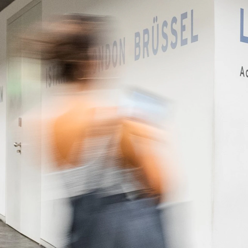 People walking through a modern, well-lit corridor with white walls displaying city names in blue. Some people are observing a display on the right wall.