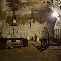 Svagt upplyst grottkammare med kors, ljus och religiösa ikoner på ett stenaltare. En träbänk och metallgrind finns också.